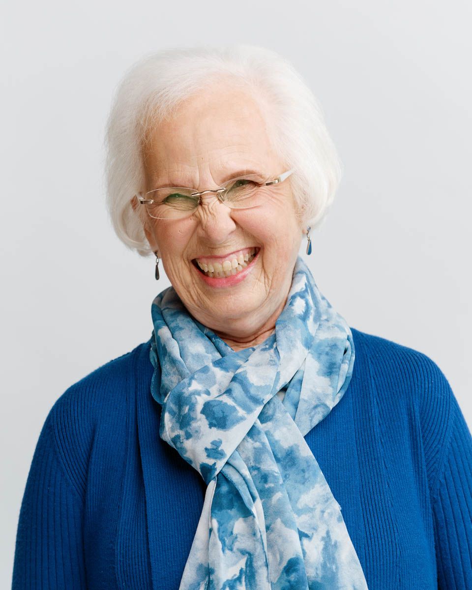 Happy elderly woman with white hair and glasses laughing, wearing a blue cardigan and floral scarf.