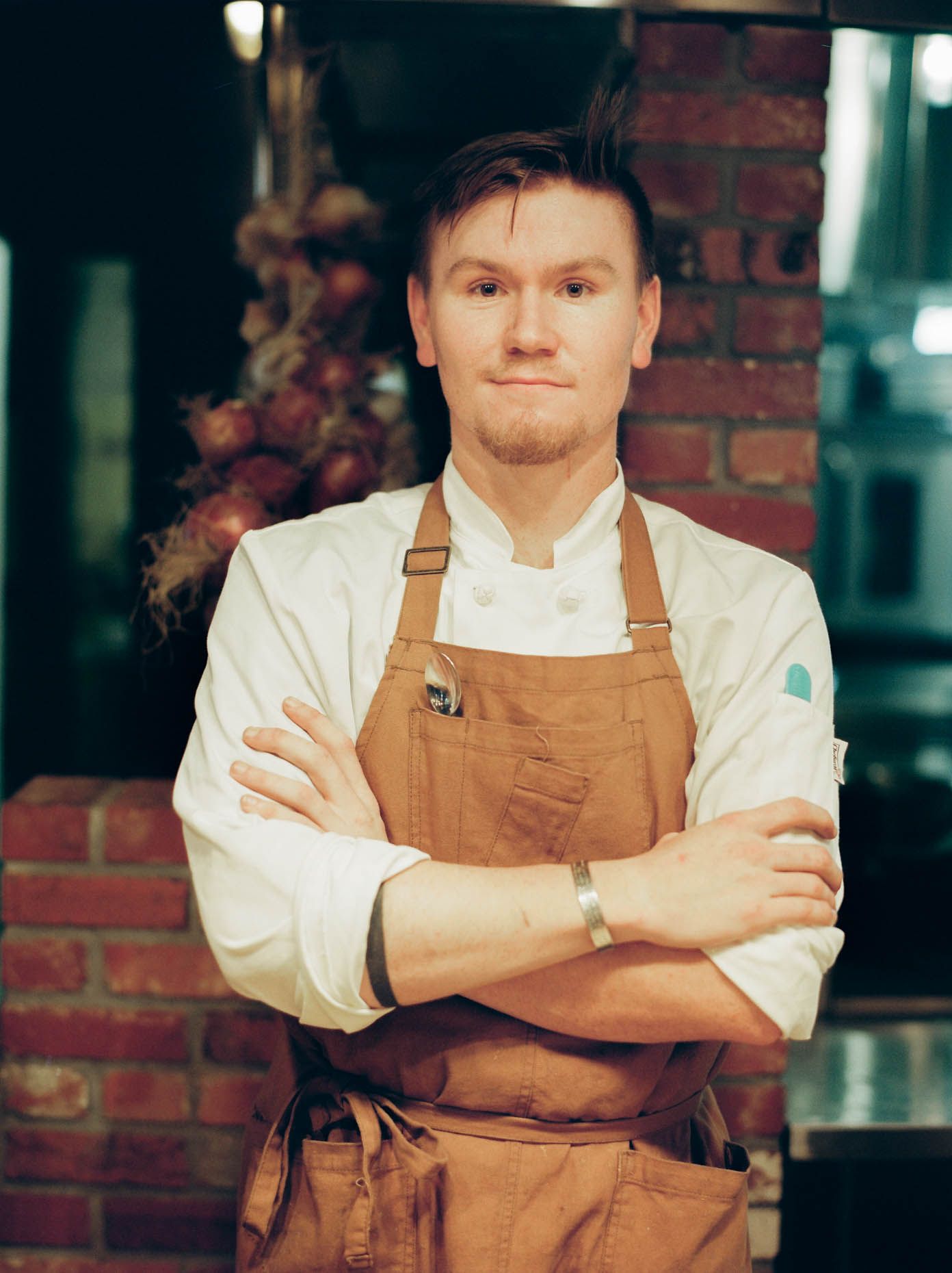 A professional chef in a white coat and brown apron stands with arms crossed in a restaurant kitchen.