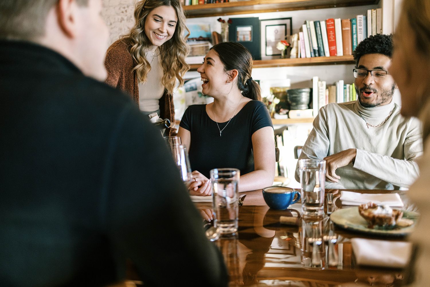 Diverse group of young adults laughing and dining together at a wooden table in a cozy restaurant.