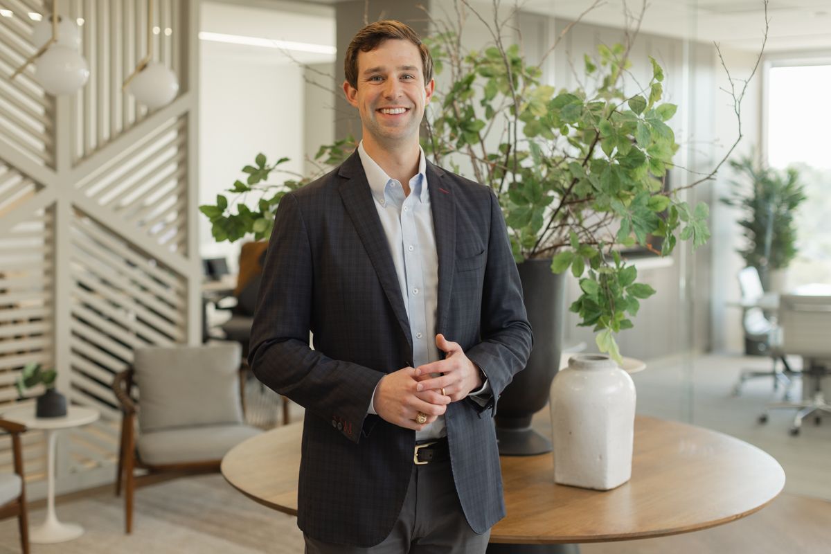 Confident businessman in blazer standing in modern office lobby with green plants and natural light.