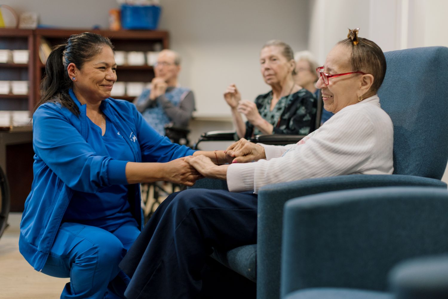 A nurse in blue scrubs holds hands with an elderly patient in a care facility, offering compassionate support.