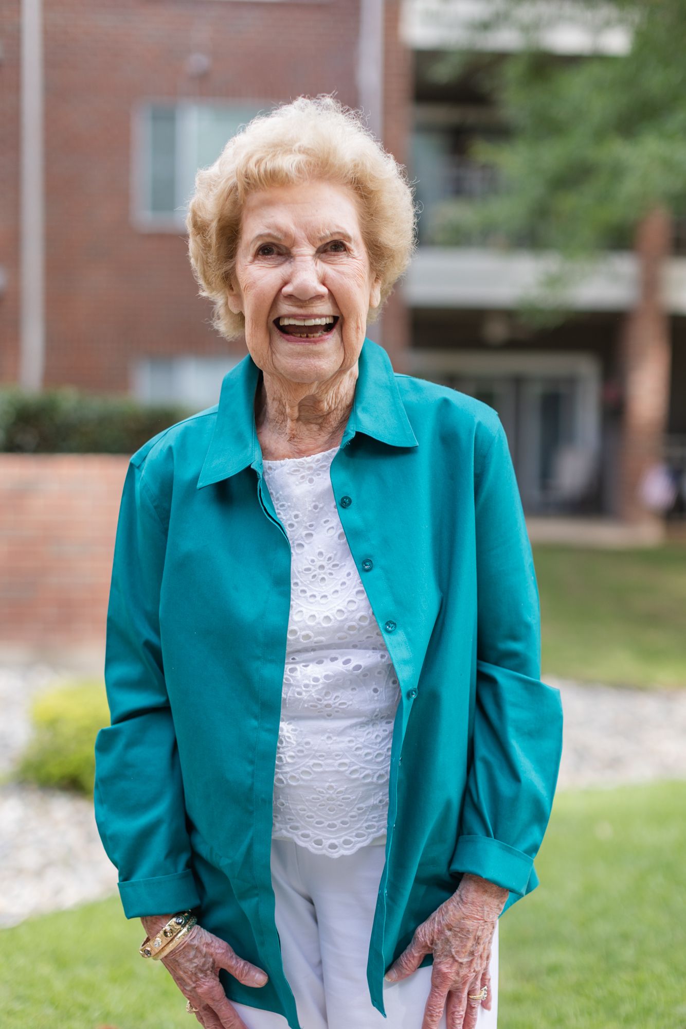 Elderly woman in teal jacket and white pants smiling outdoors near a brick building and green lawn.