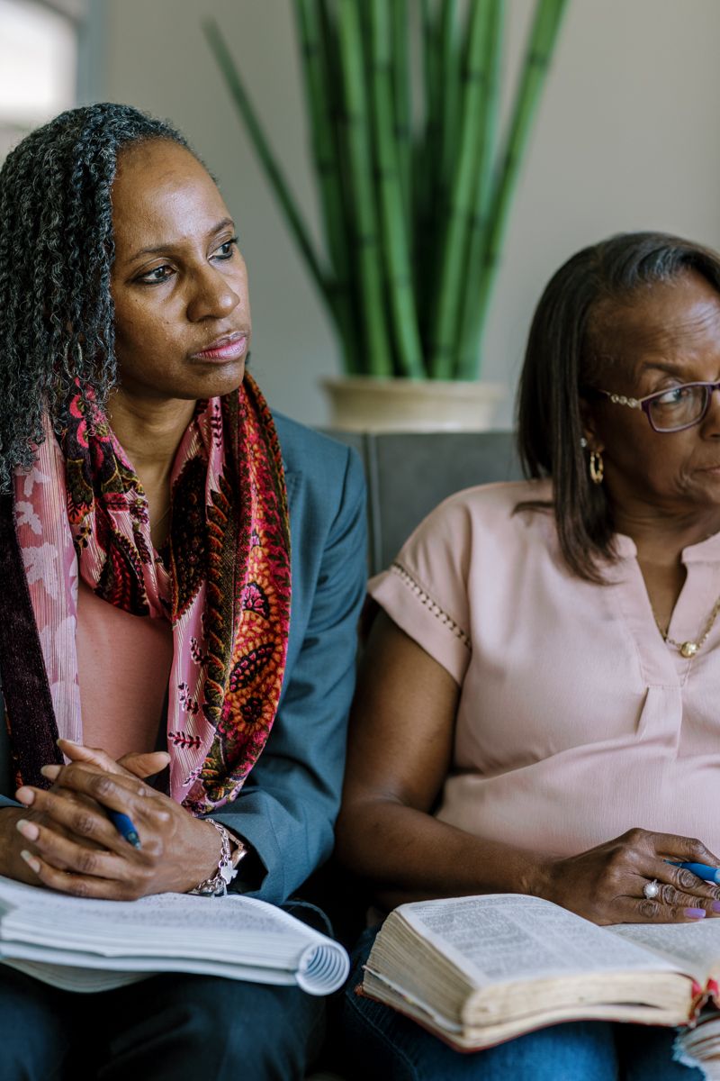 Two Black women sitting together studying open Bibles, one wearing a colorful scarf and blazer, the other in a pink blouse.