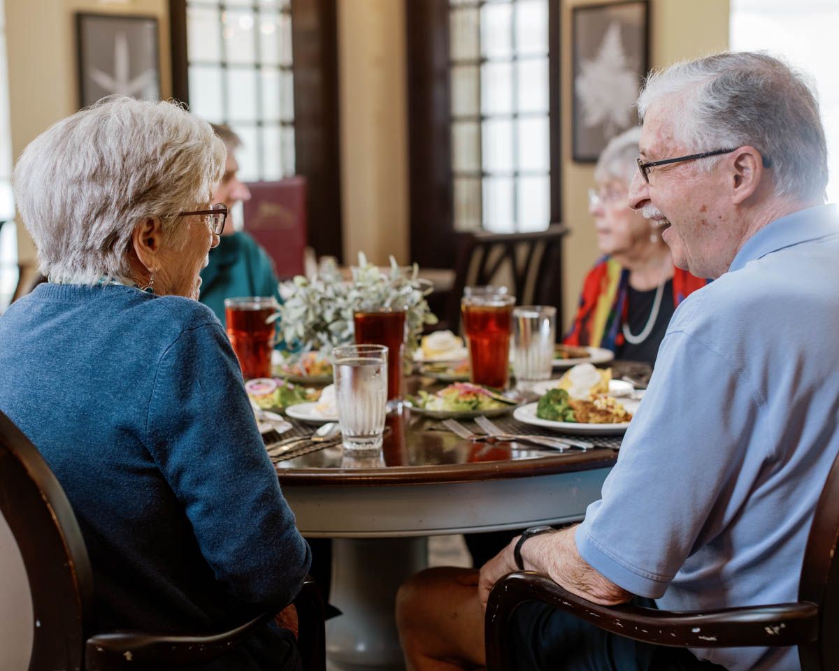 Elderly group enjoying a meal together at a round dining table with drinks and food in a warm restaurant setting.