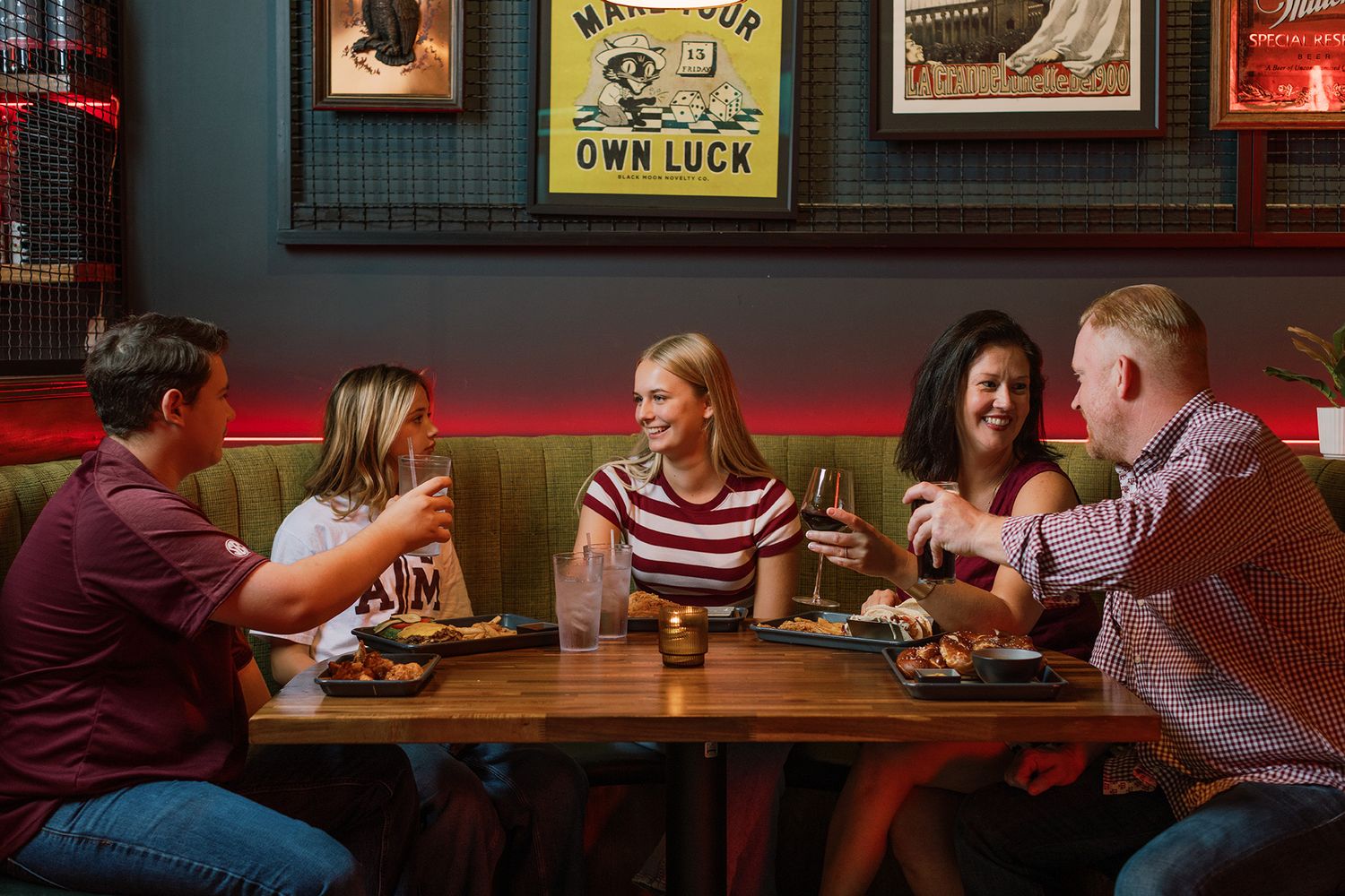 Group of five friends laughing and toasting drinks over dinner at a restaurant.