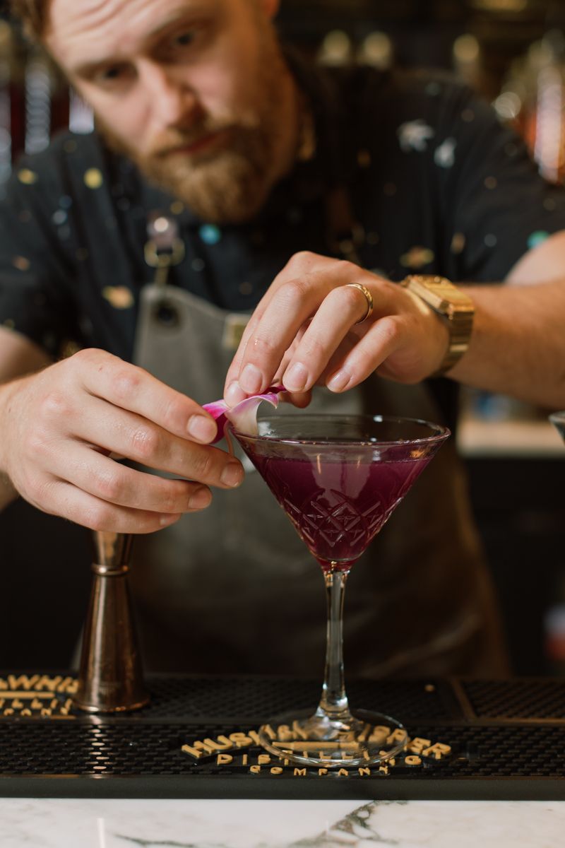 Bearded bartender garnishing a purple cocktail in a martini glass at a bar.