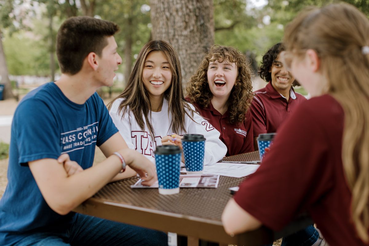 Five college students laughing and socializing at an outdoor picnic table with Texas A&M cups on campus.