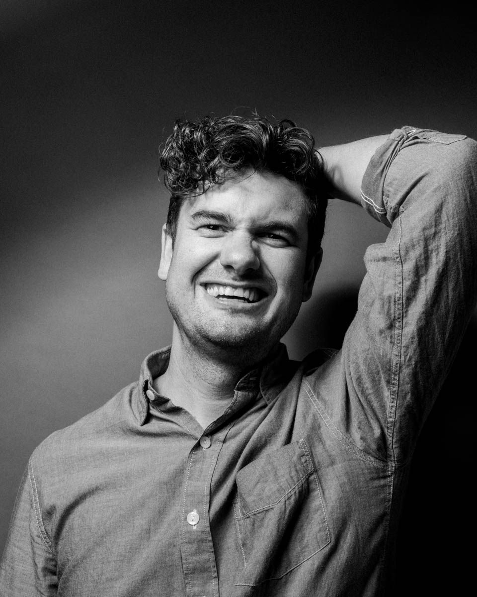 Black and white photo of a laughing young man with curly hair posing with hand behind head.