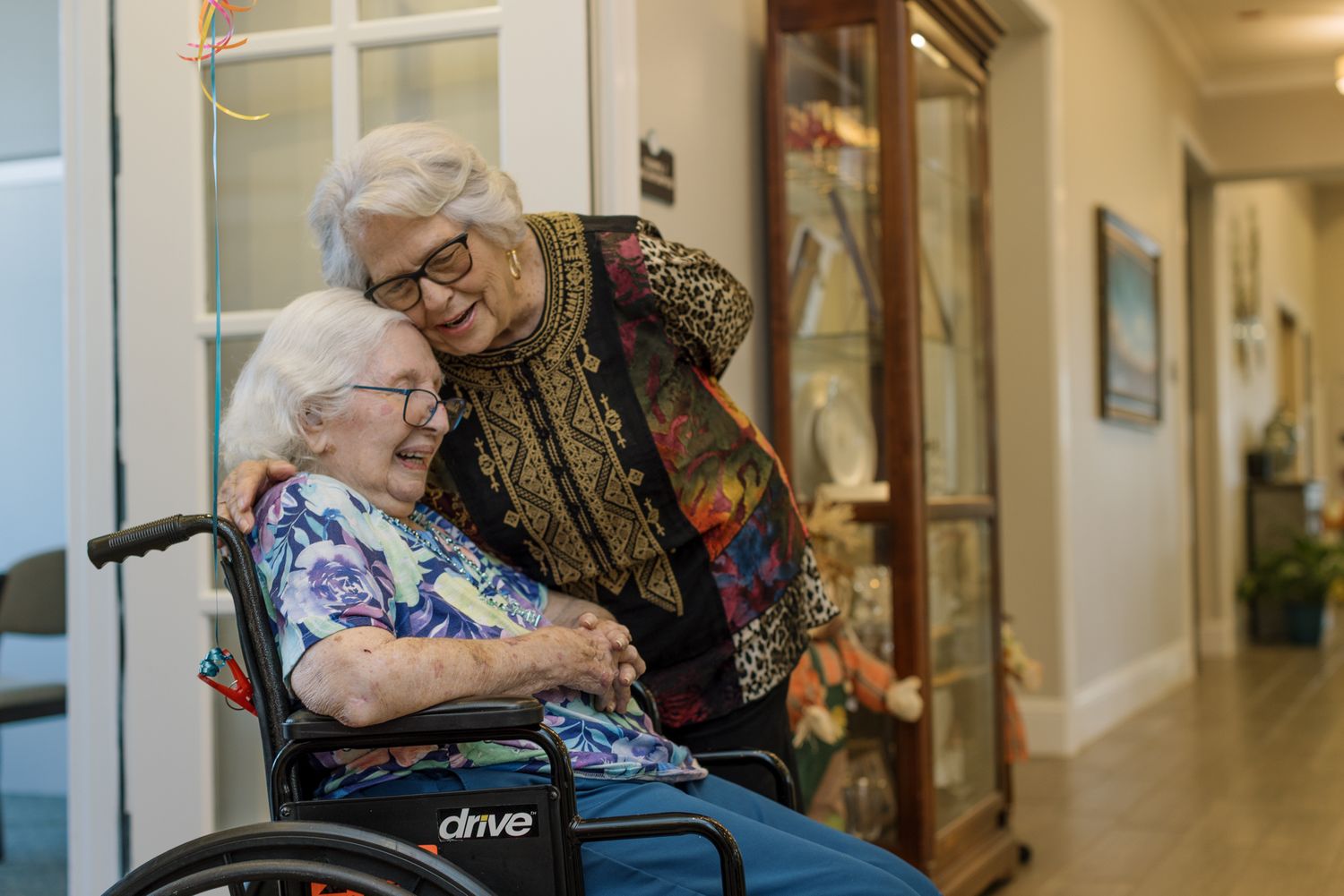 An elderly woman in a wheelchair shares a warm embrace with an older female caregiver in a senior living facility hallway.