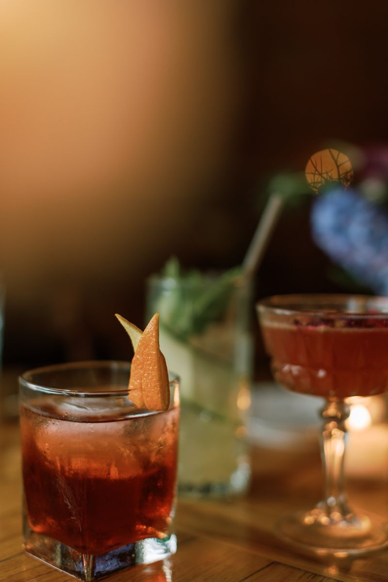 Three craft cocktails on a wooden bar, featuring a red drink with a dried citrus garnish in a rocks glass.