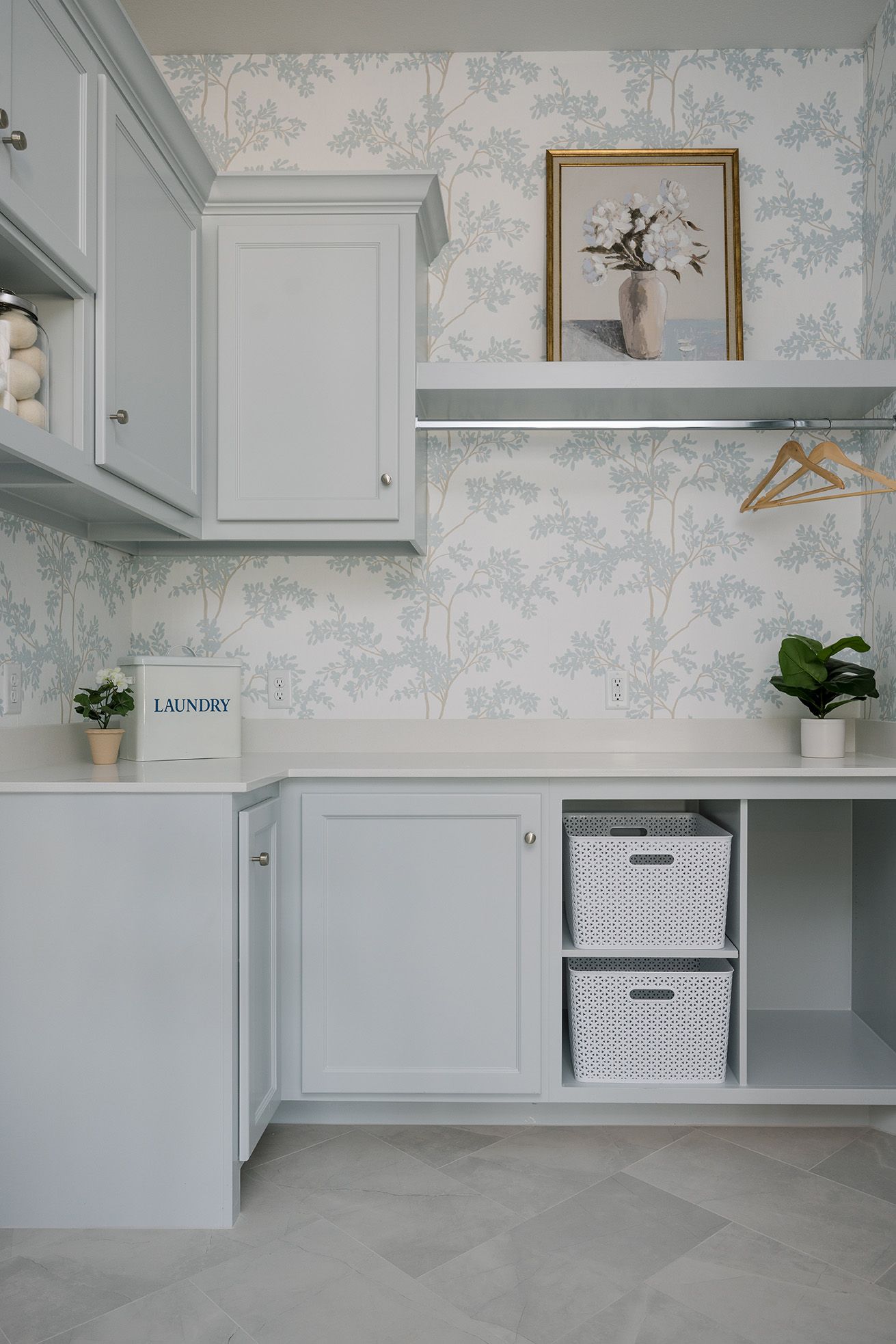 Light gray laundry room with floral wallpaper, open shelving, wicker storage baskets, and white cabinetry.