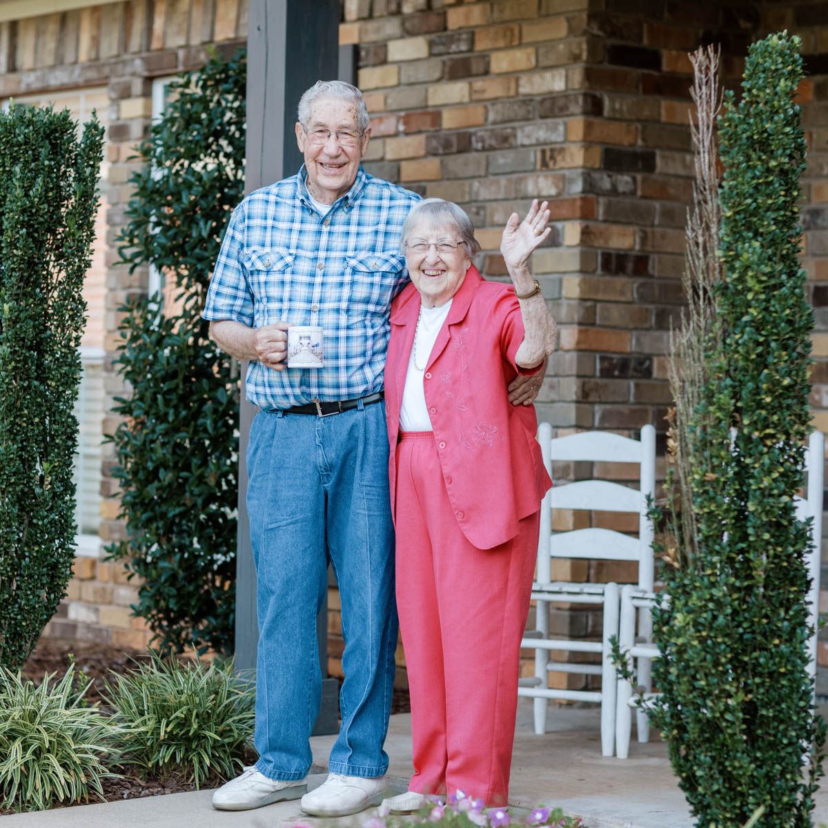 Happy elderly couple standing on a brick home porch, smiling and waving at the camera outdoors.