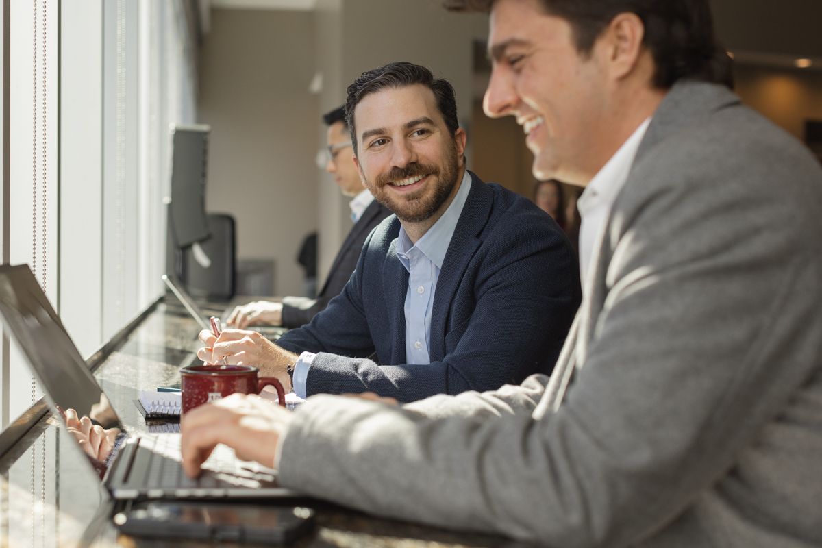 Three businessmen smiling and working on laptops together at a counter in a modern office setting.