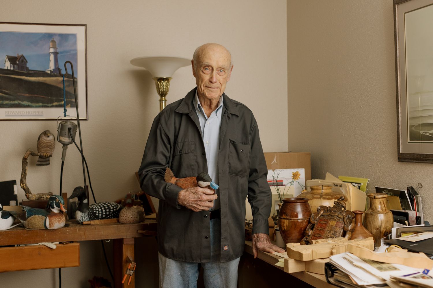 Elderly man standing in his workshop surrounded by wooden crafts and collectibles.