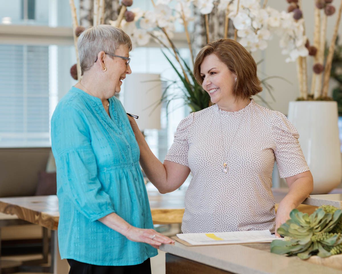 Two women smiling and talking together in a bright, modern interior setting with decorative plants nearby.