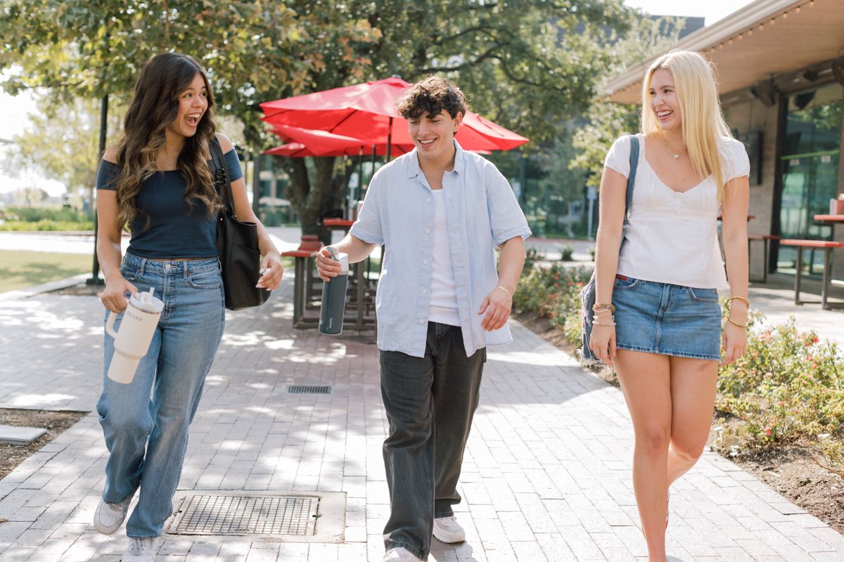 Three smiling college students walk together on a sunny campus sidewalk outdoors.