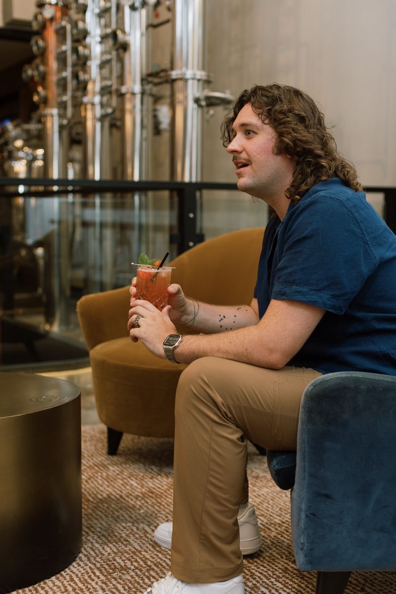 Young man with curly hair sitting in a lounge chair holding a cocktail near distillery equipment.