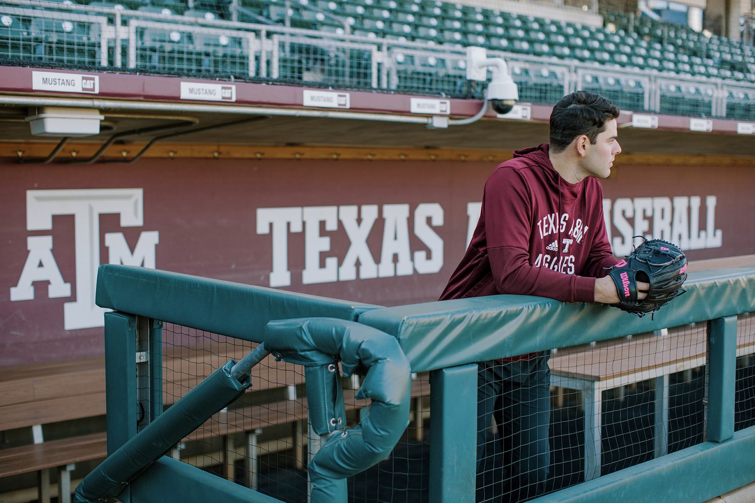 Texas A&M baseball player in maroon hoodie leaning on dugout fence at stadium.