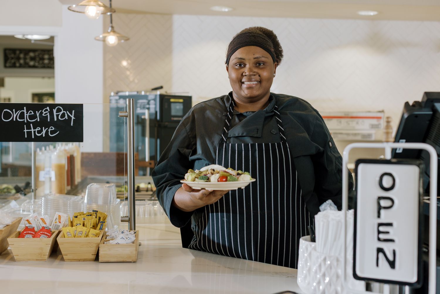 Friendly female chef in black apron presents a fresh salad plate at an open deli counter.