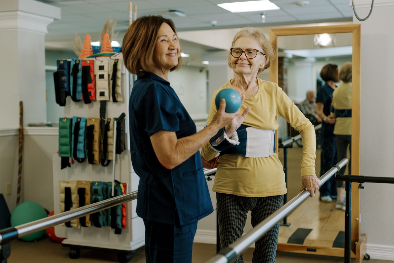 A physical therapist assists an elderly woman with balance exercises using parallel bars in a rehabilitation facility.