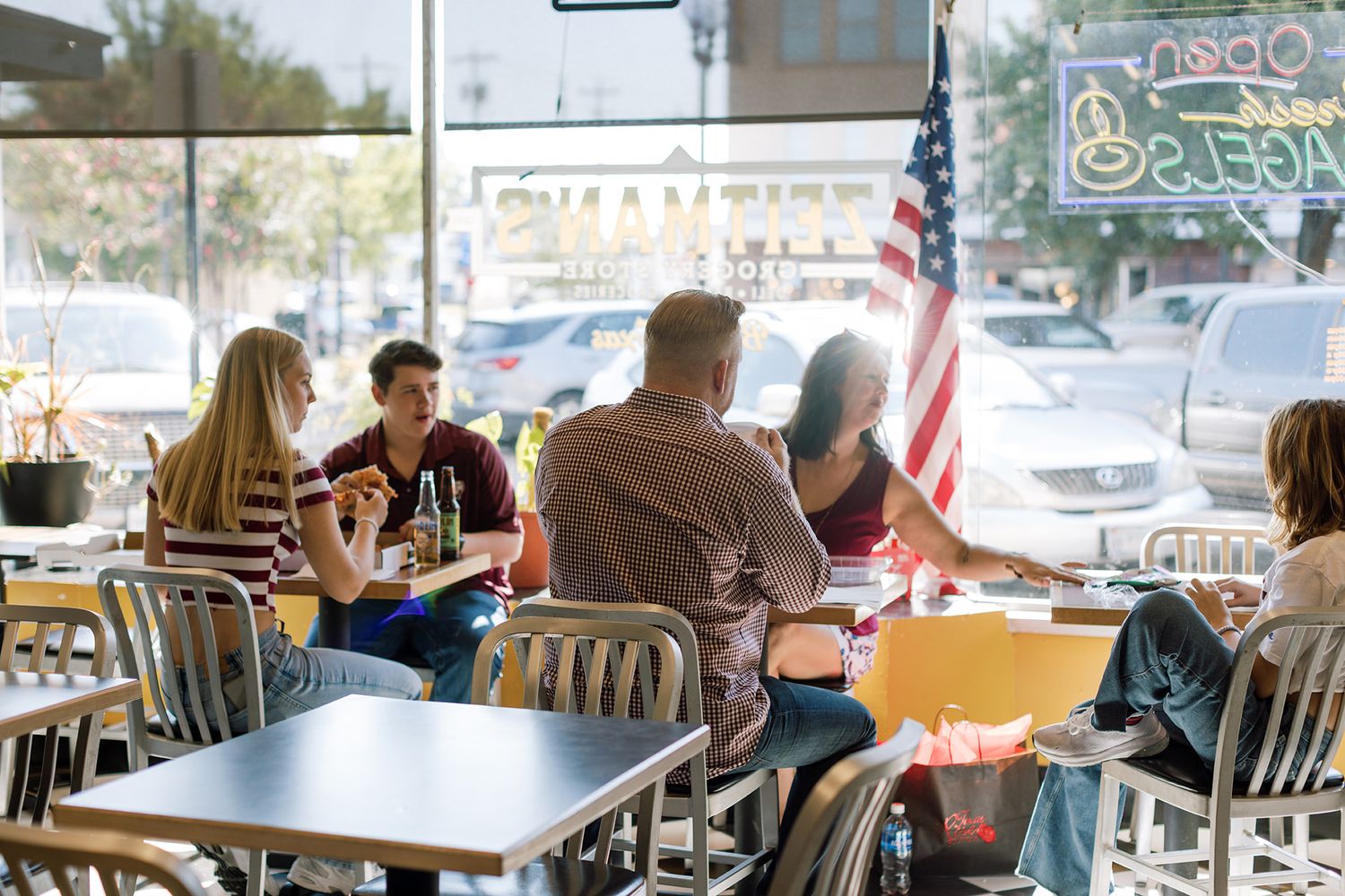 Group of friends dining at a casual open-front restaurant with an American flag and neon signs in background.