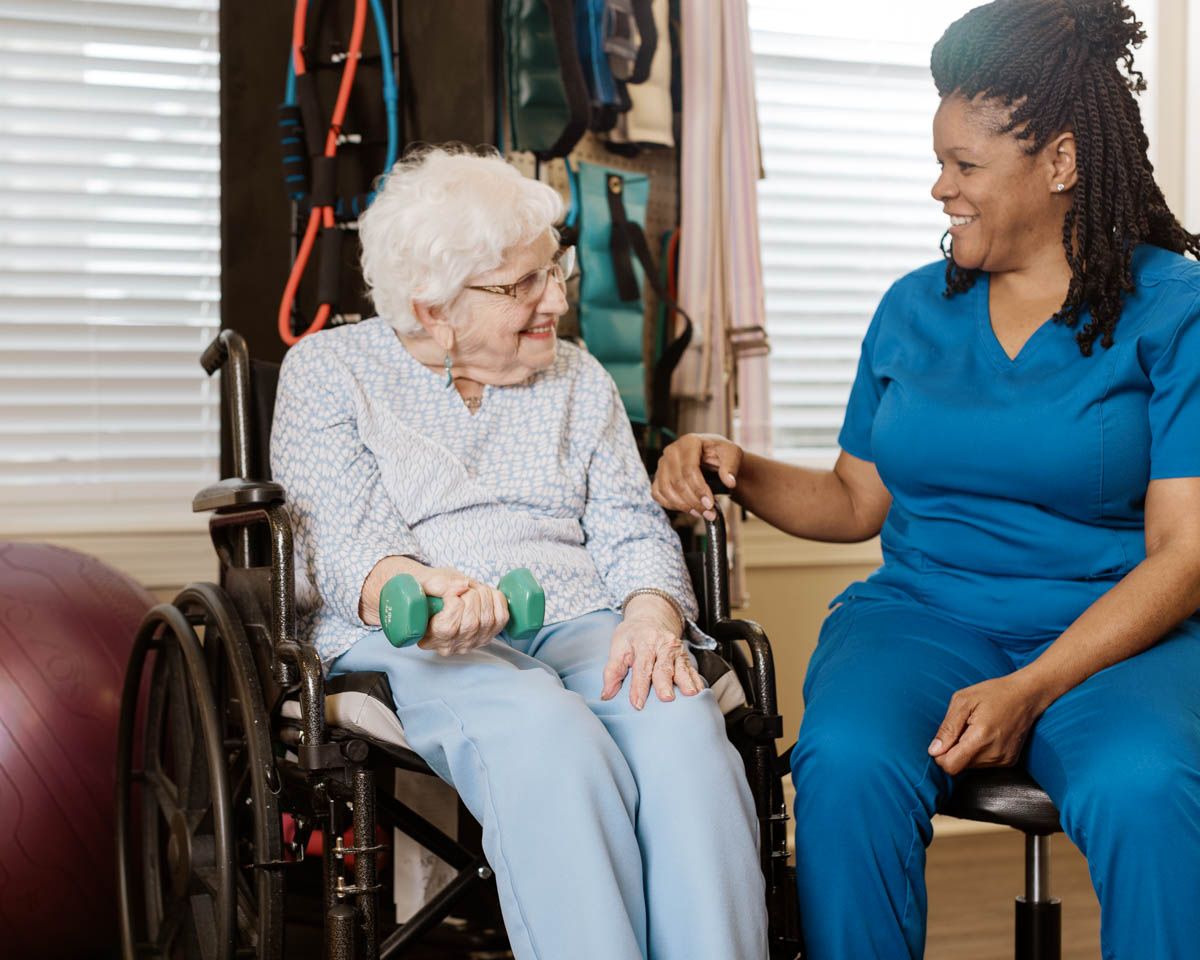 Elderly woman in wheelchair doing dumbbell exercises with a smiling female nurse in blue scrubs at a care facility.
