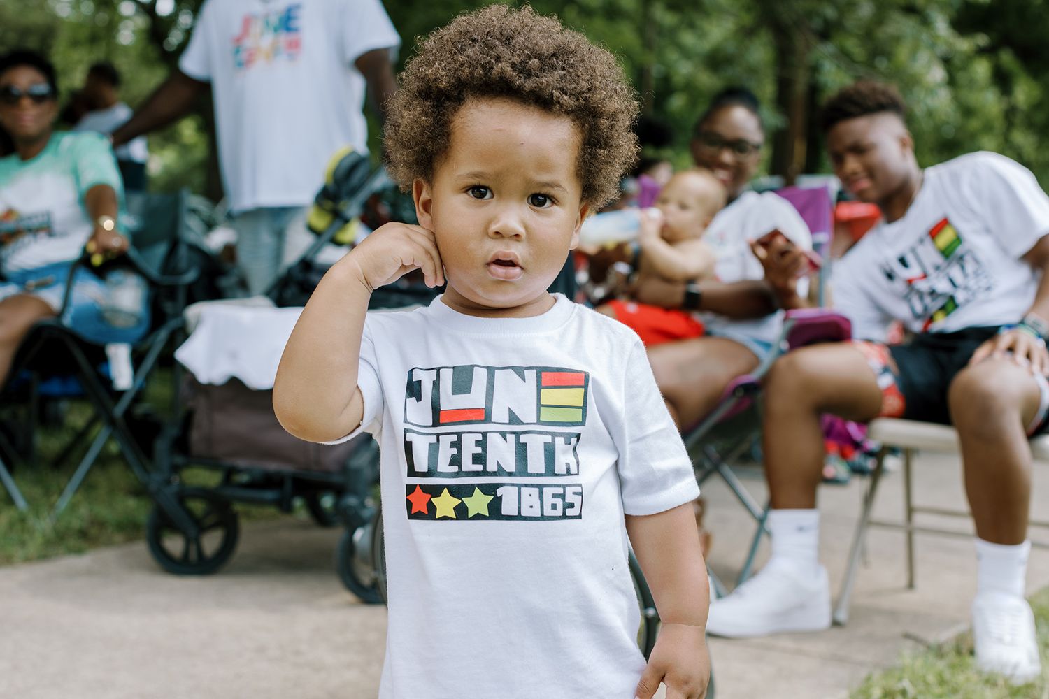 Toddler wearing a white Juneteenth 1865 t-shirt stands confidently at an outdoor celebration.