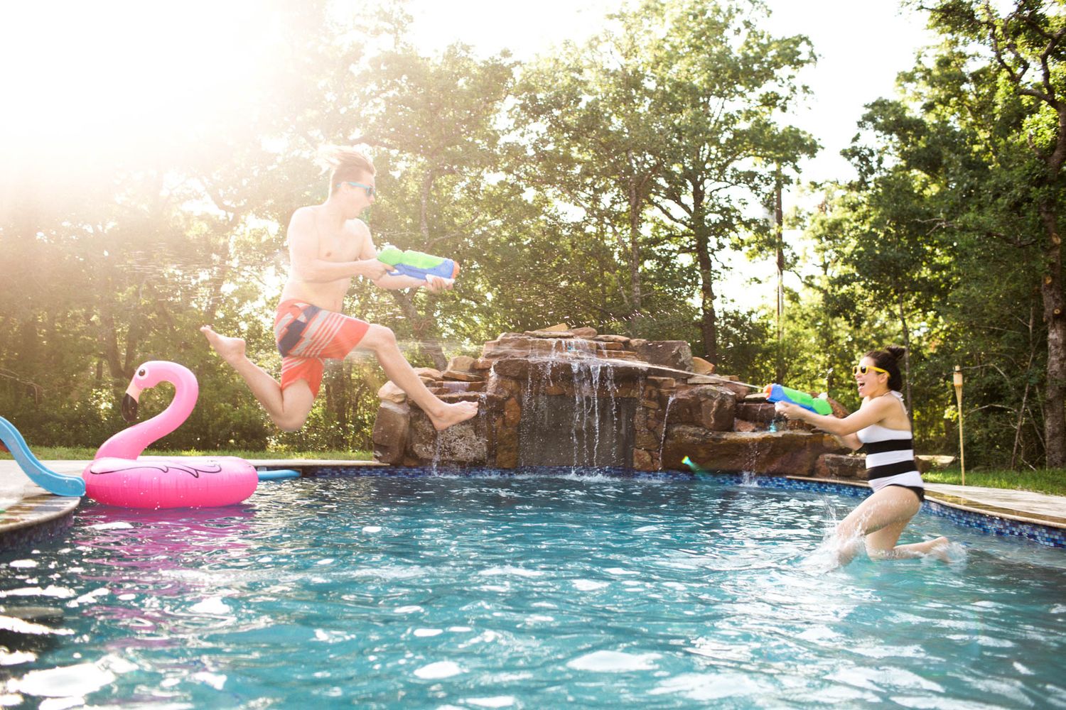 Two people having a water gun fight in a backyard pool with a waterfall feature and pink flamingo float.