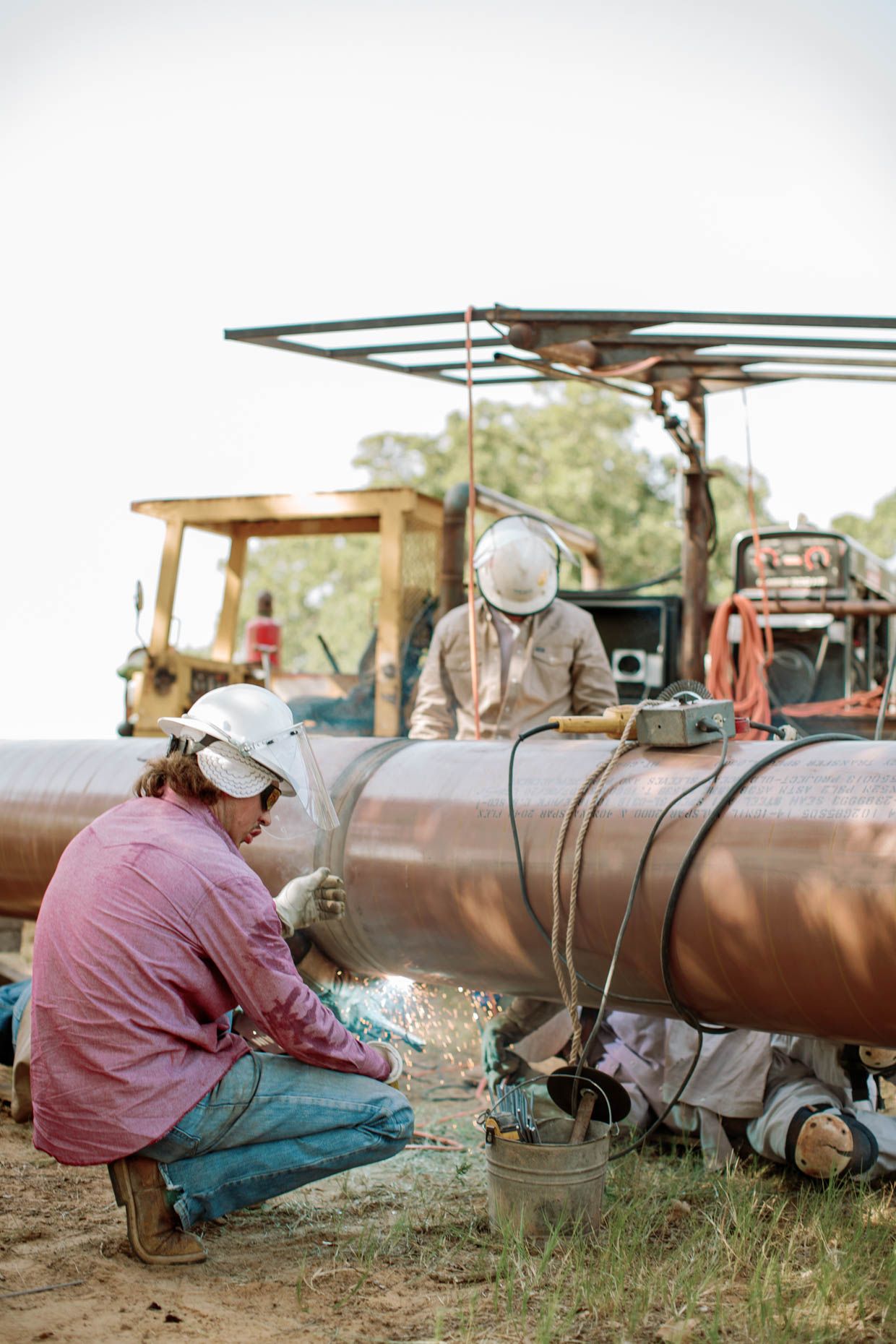 Two workers in hard hats welding a large industrial pipeline outdoors near heavy machinery.