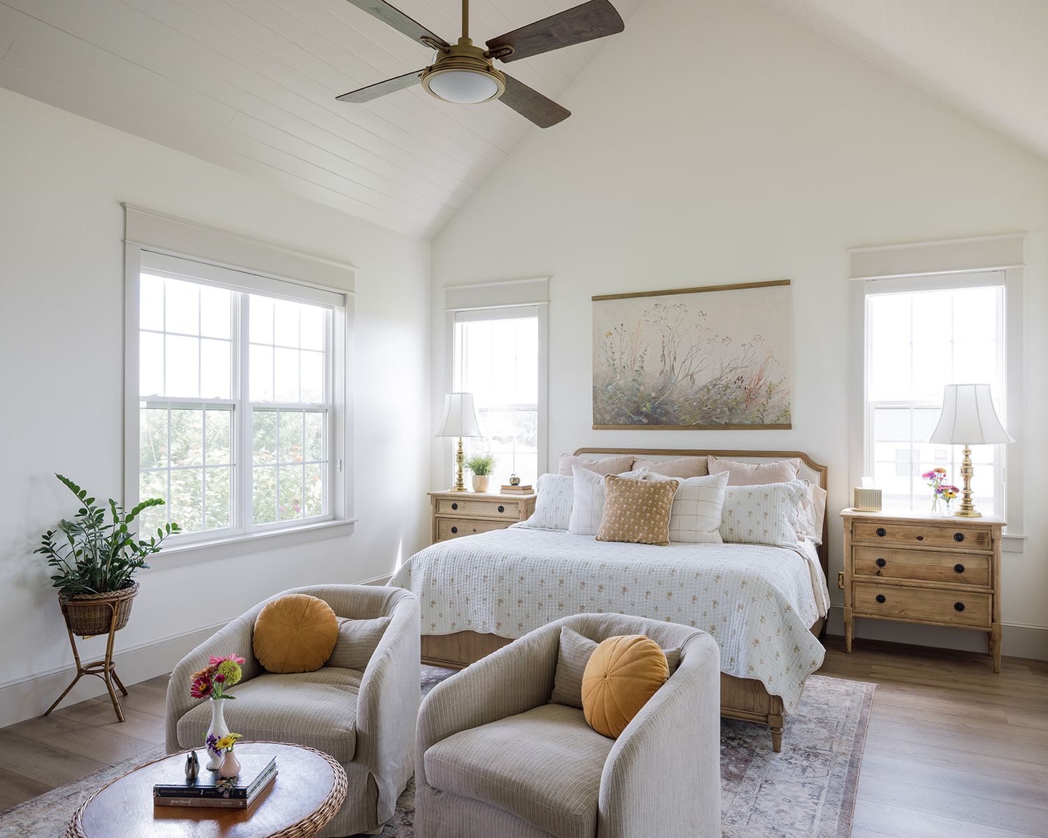 Bright master bedroom with vaulted ceiling, neutral linen chairs, wood dresser, and ceiling fan.