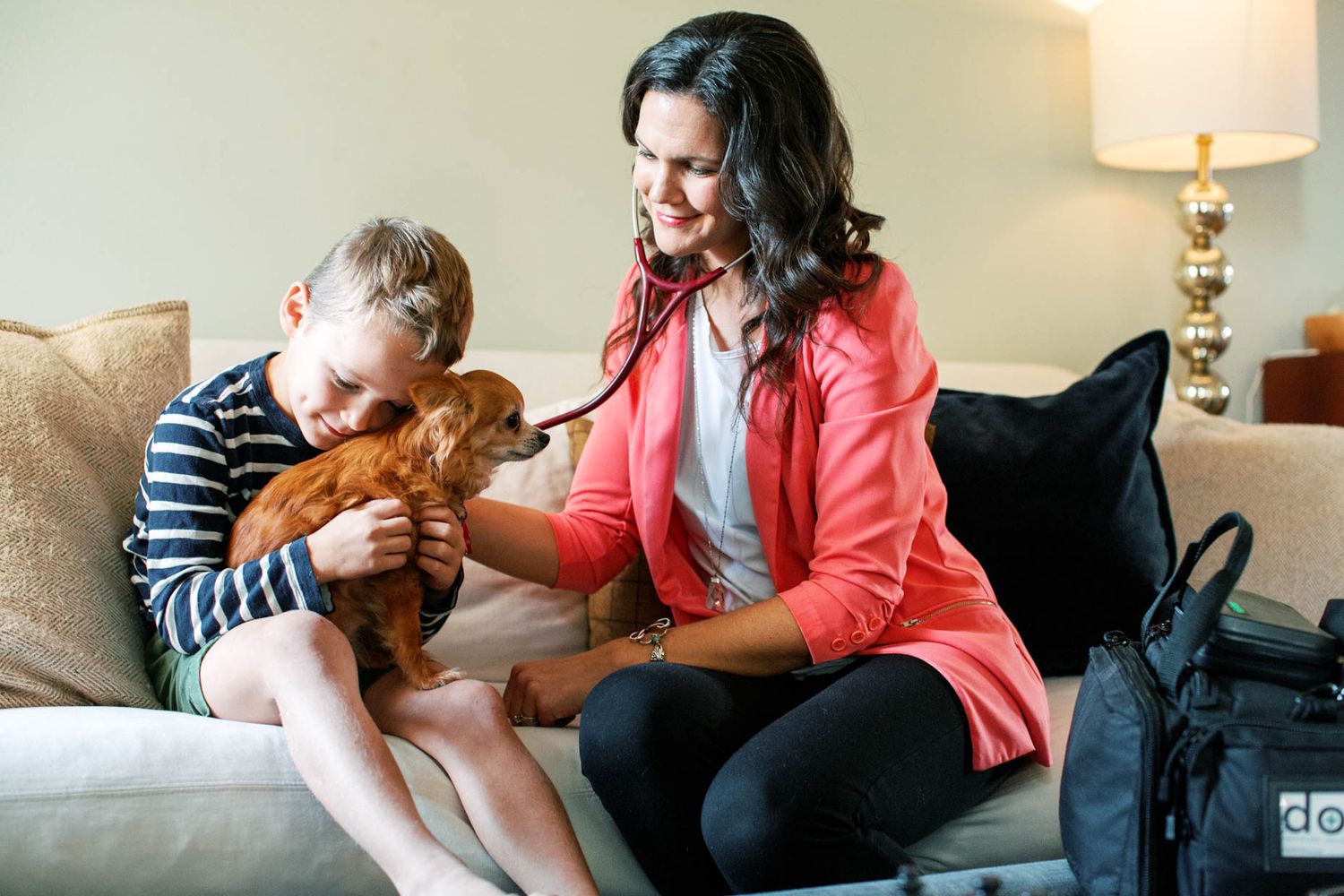 A woman in pink uses a stethoscope on a stuffed animal toy while a young boy sits beside her on a sofa.