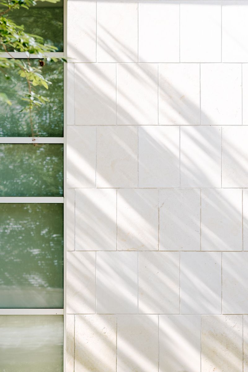 Sunlight casts diagonal shadows on a white tiled wall beside large windows reflecting green trees outside.