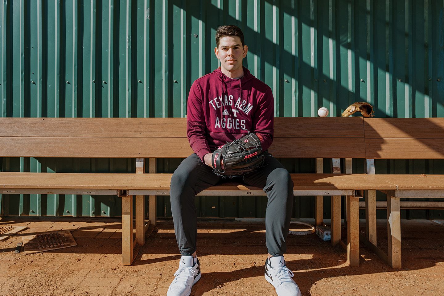 Texas A&M Aggies baseball player sitting on bench holding glove and ball.