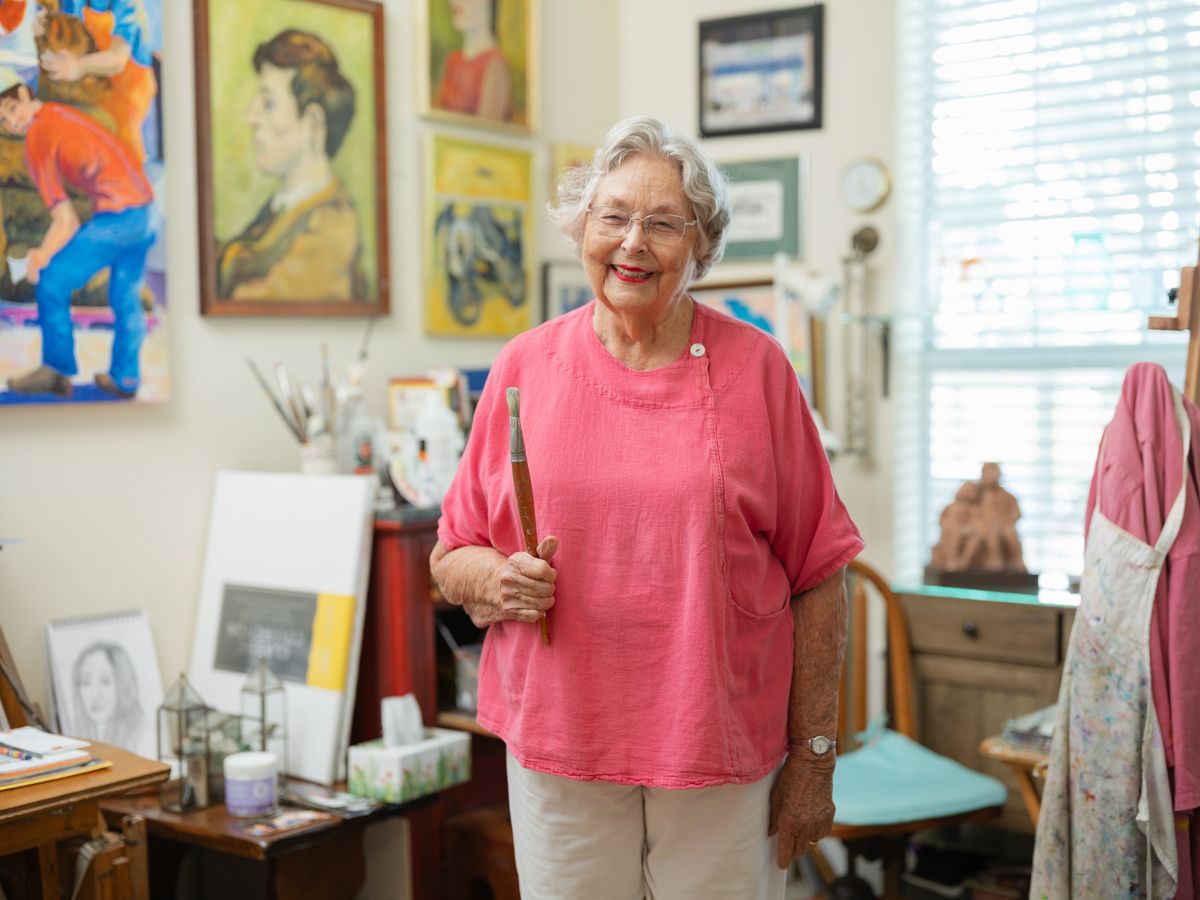 Elderly woman artist smiling in her art studio, holding a paintbrush, surrounded by colorful paintings and artwork.