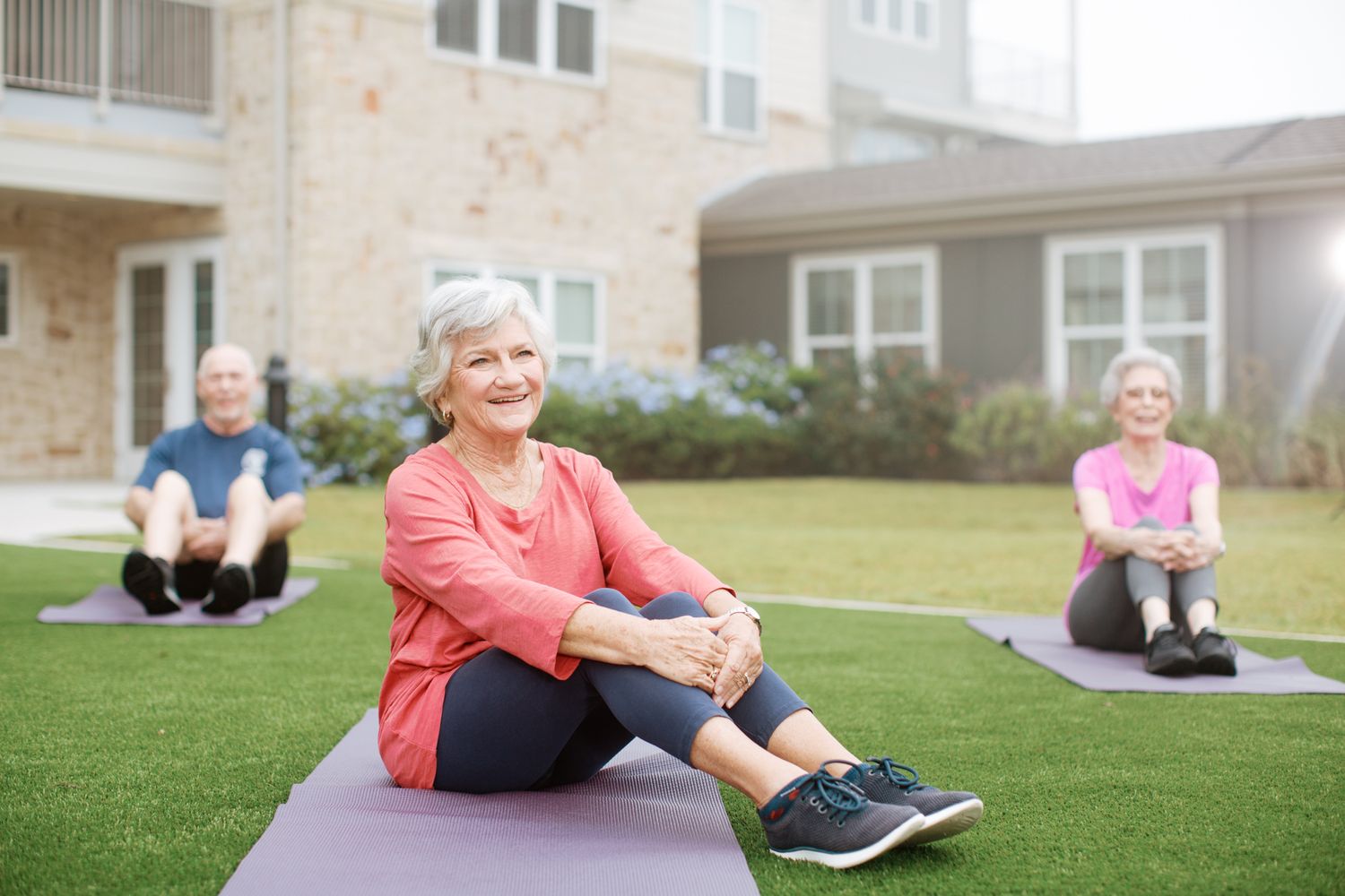 A smiling senior woman in red sits on a yoga mat outdoors during a group fitness class on green grass.