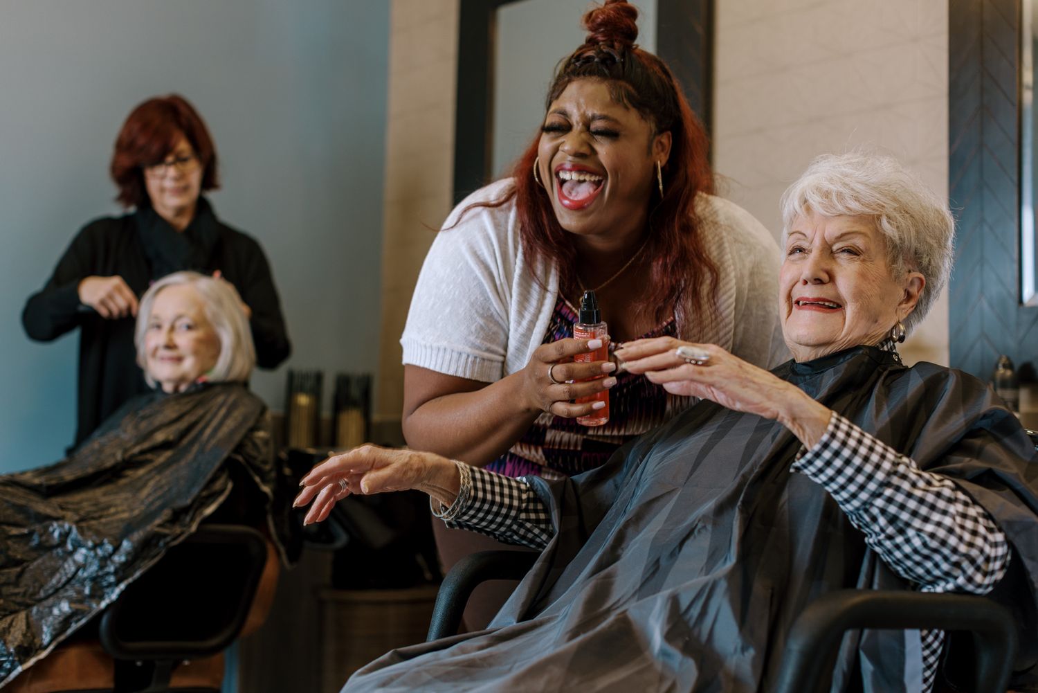 A joyful hairstylist laughs with an elderly woman in a salon chair while another stylist works in the background.