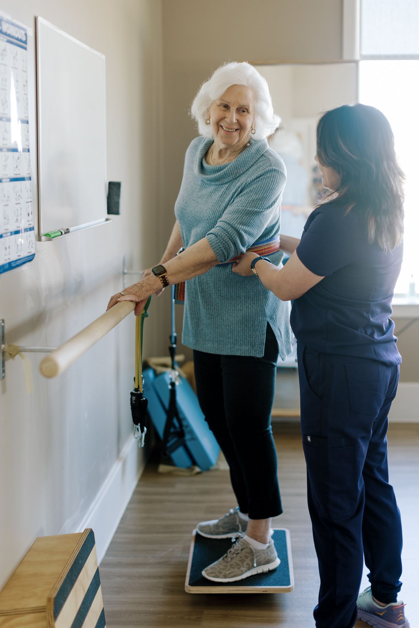 Elderly woman smiling during physical therapy session, balancing on a board while assisted by a healthcare professional.