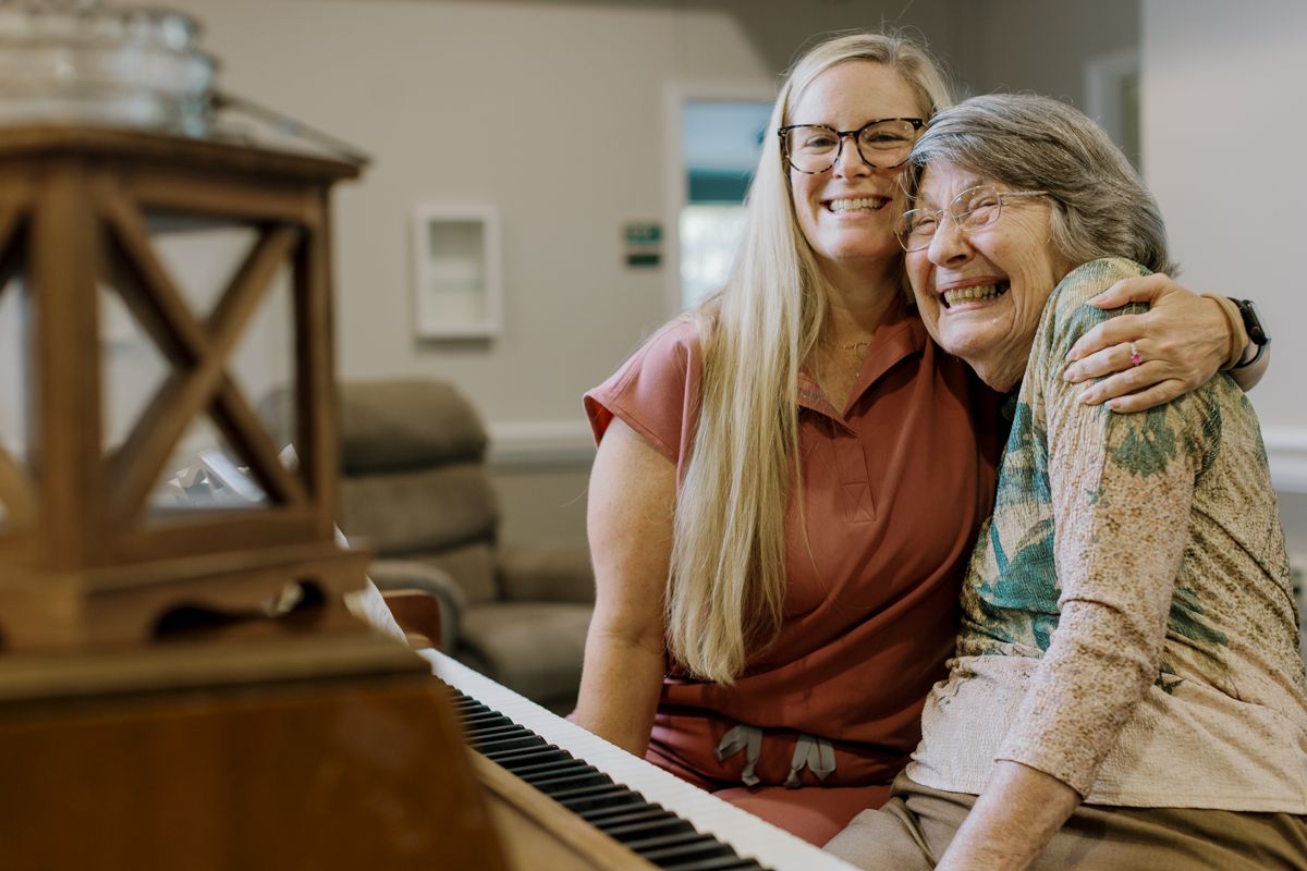 A young woman and elderly woman share a warm hug while sitting at a piano together.