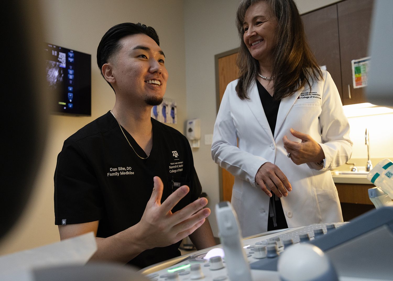Two medical professionals in a clinical setting engage in a friendly conversation near equipment.