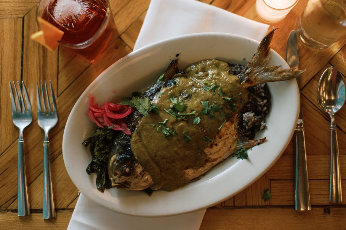 Whole roasted fish served in a white oval dish with greens and pickled onions on a wooden table.
