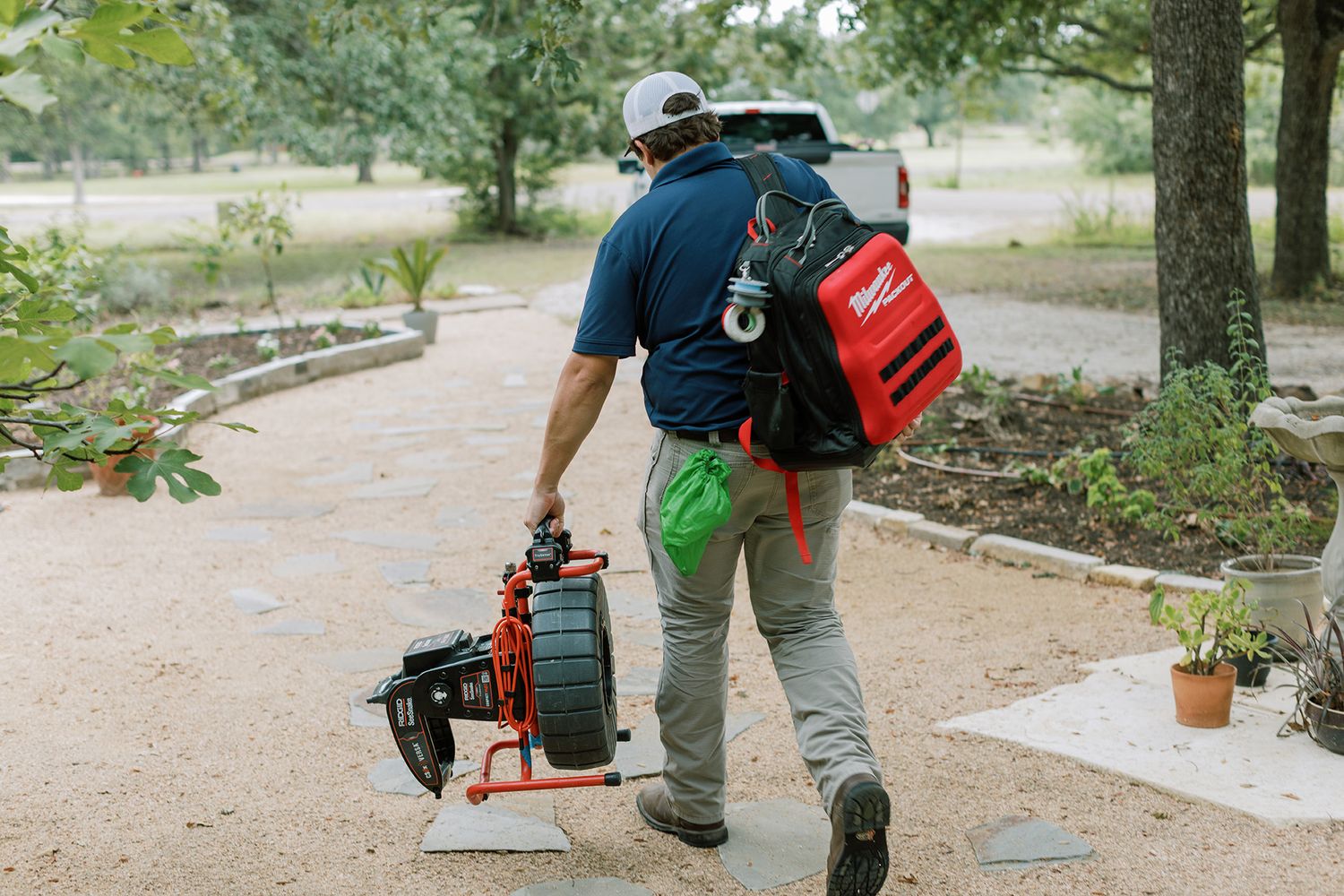 Plumber carrying Milwaukee tools and drain cleaning equipment along a garden path toward a residential property.