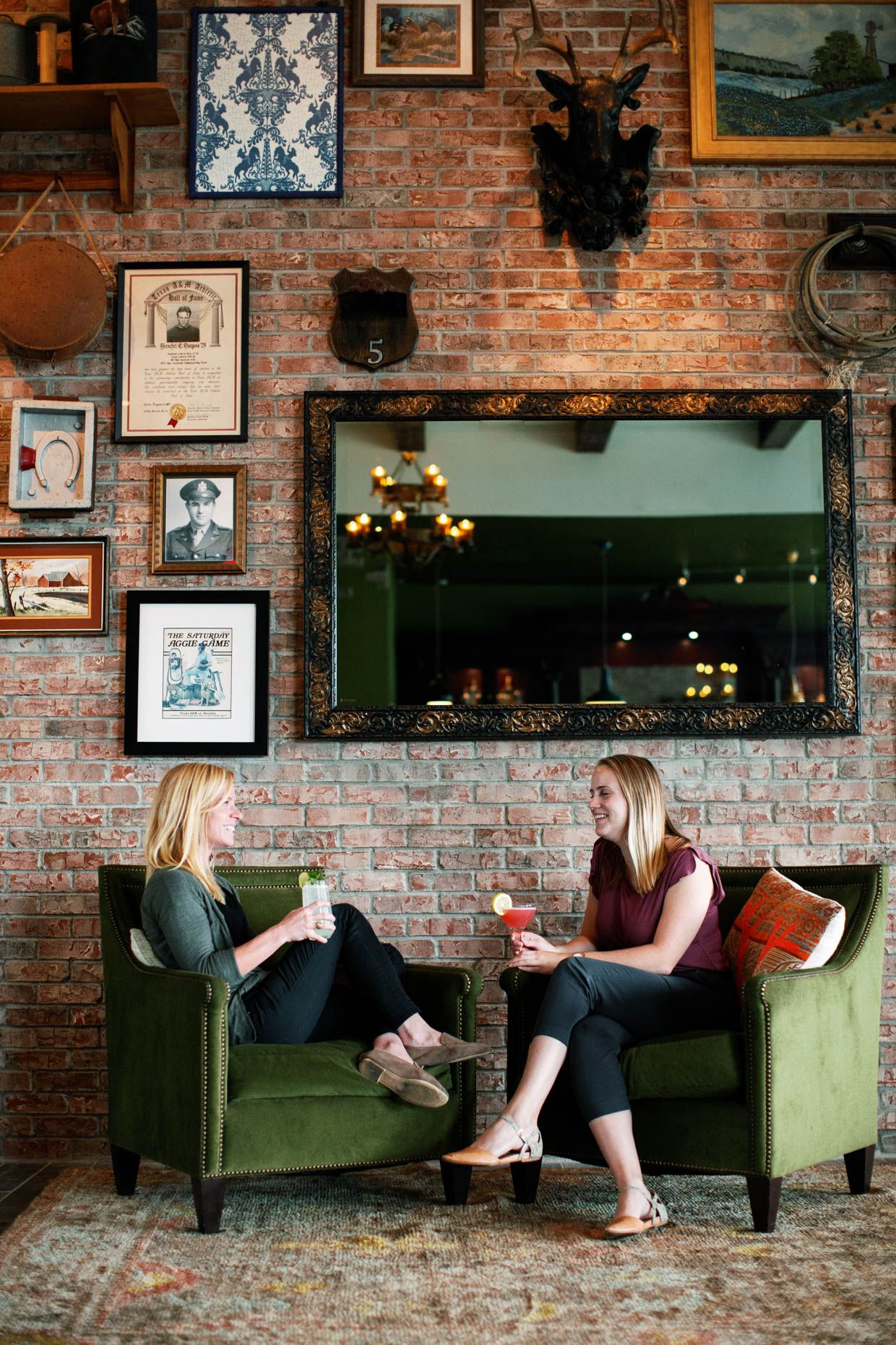 Two women enjoy cocktails in green velvet chairs against a rustic brick wall adorned with vintage frames and a large mirror.