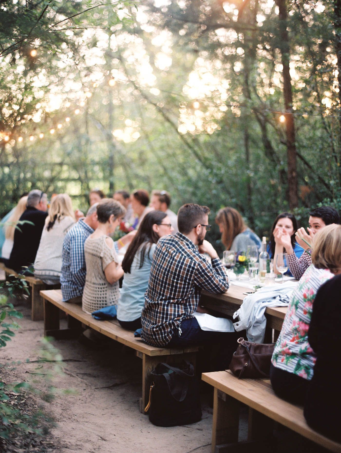 Guests dining outdoors at long wooden tables surrounded by trees with string lights at a forest dinner event.