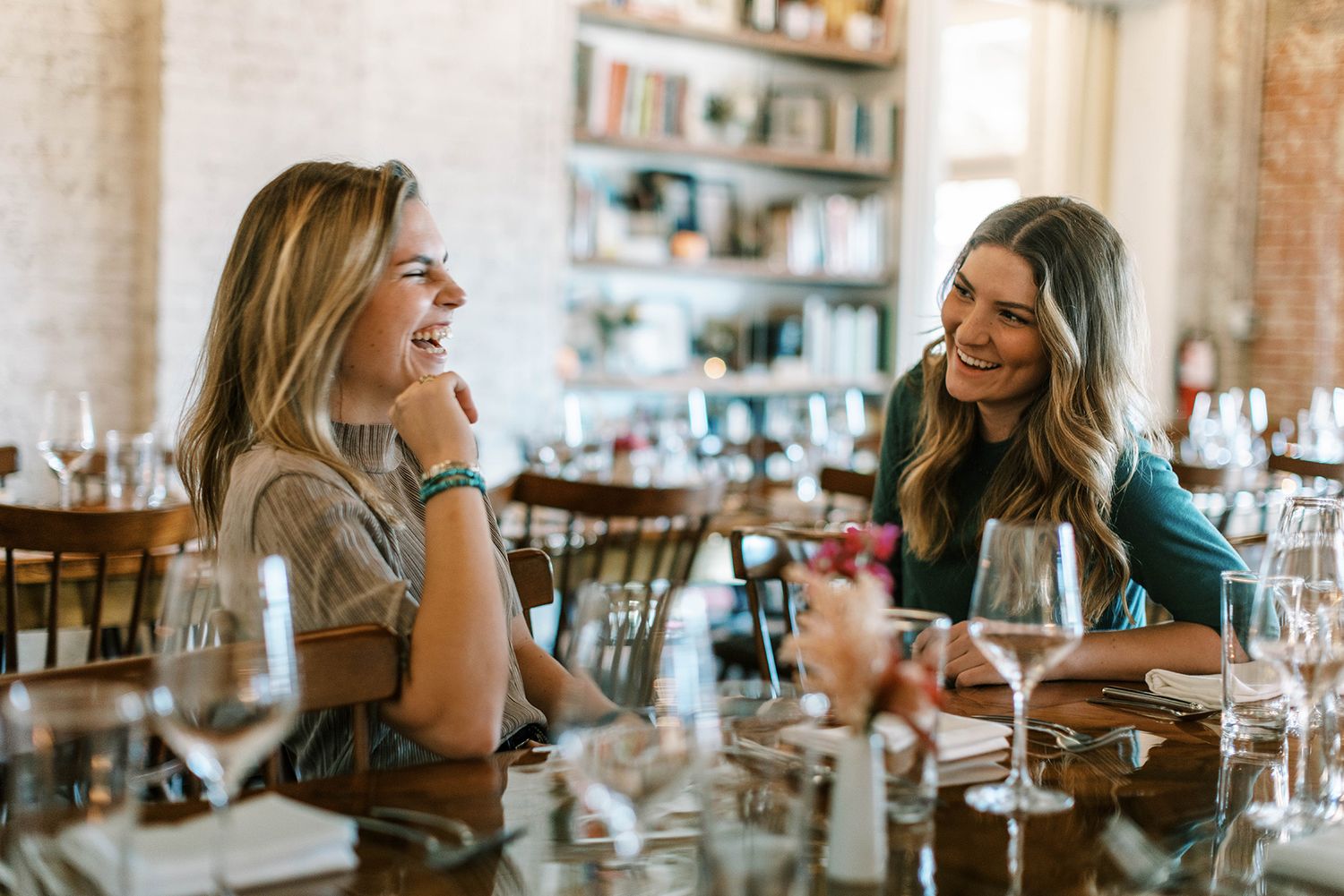 Two women laughing and enjoying conversation at a restaurant table with wine glasses in a bright interior.