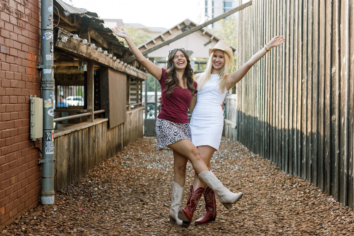 Two young women celebrating with arms raised in a rustic outdoor alleyway setting.