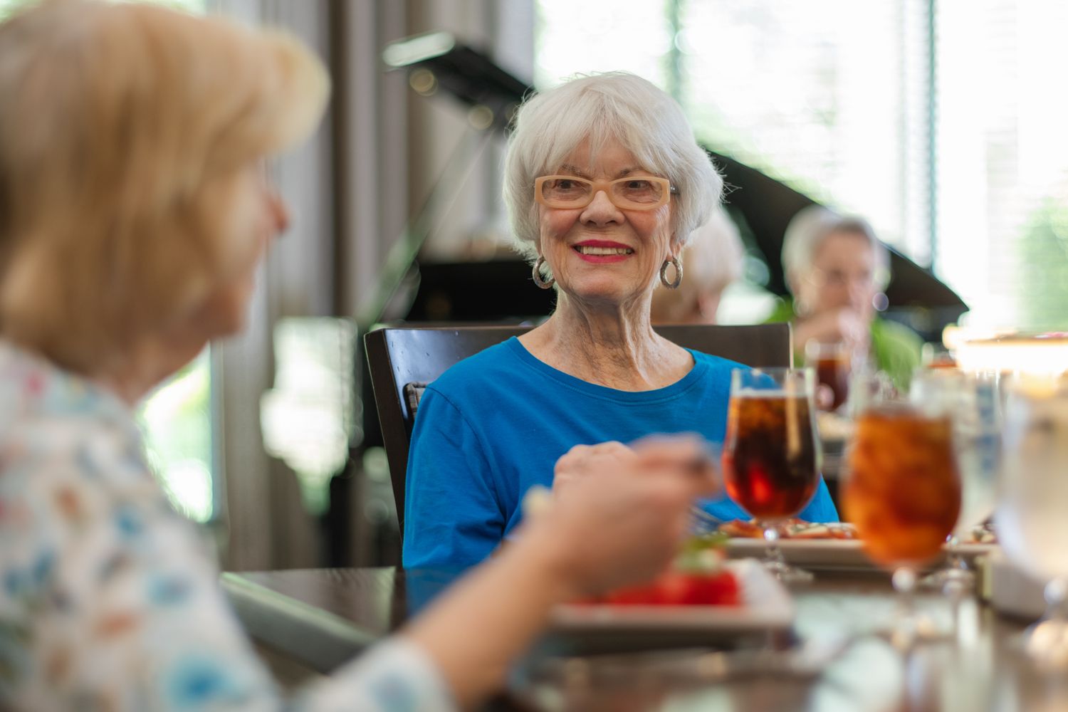 Senior women smiling and socializing over lunch at a bright restaurant, enjoying iced tea and fresh fruit together.