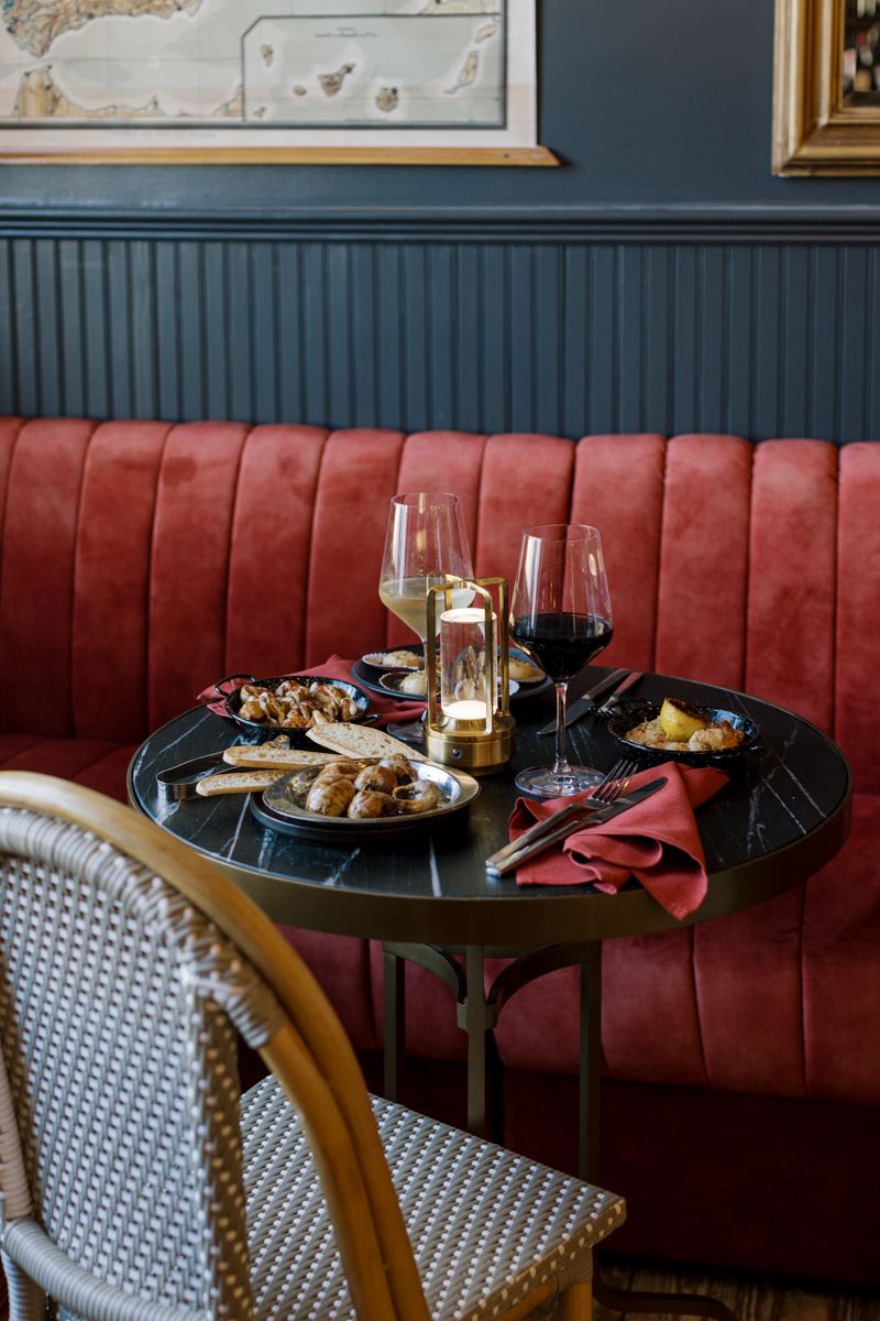 Elegant restaurant table set with wine glasses, candle, and appetizers beside a red velvet banquette in a moody dining room.