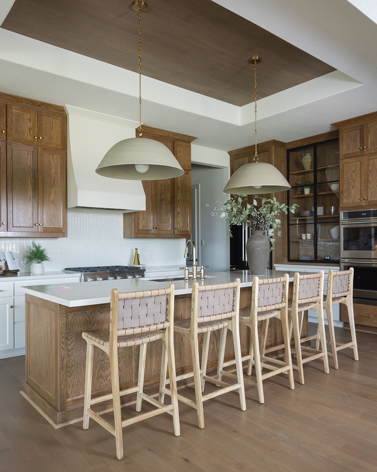 Spacious kitchen with rich wood cabinetry, large white island, woven counter stools, and sage green pendant lights on tray ceiling.
