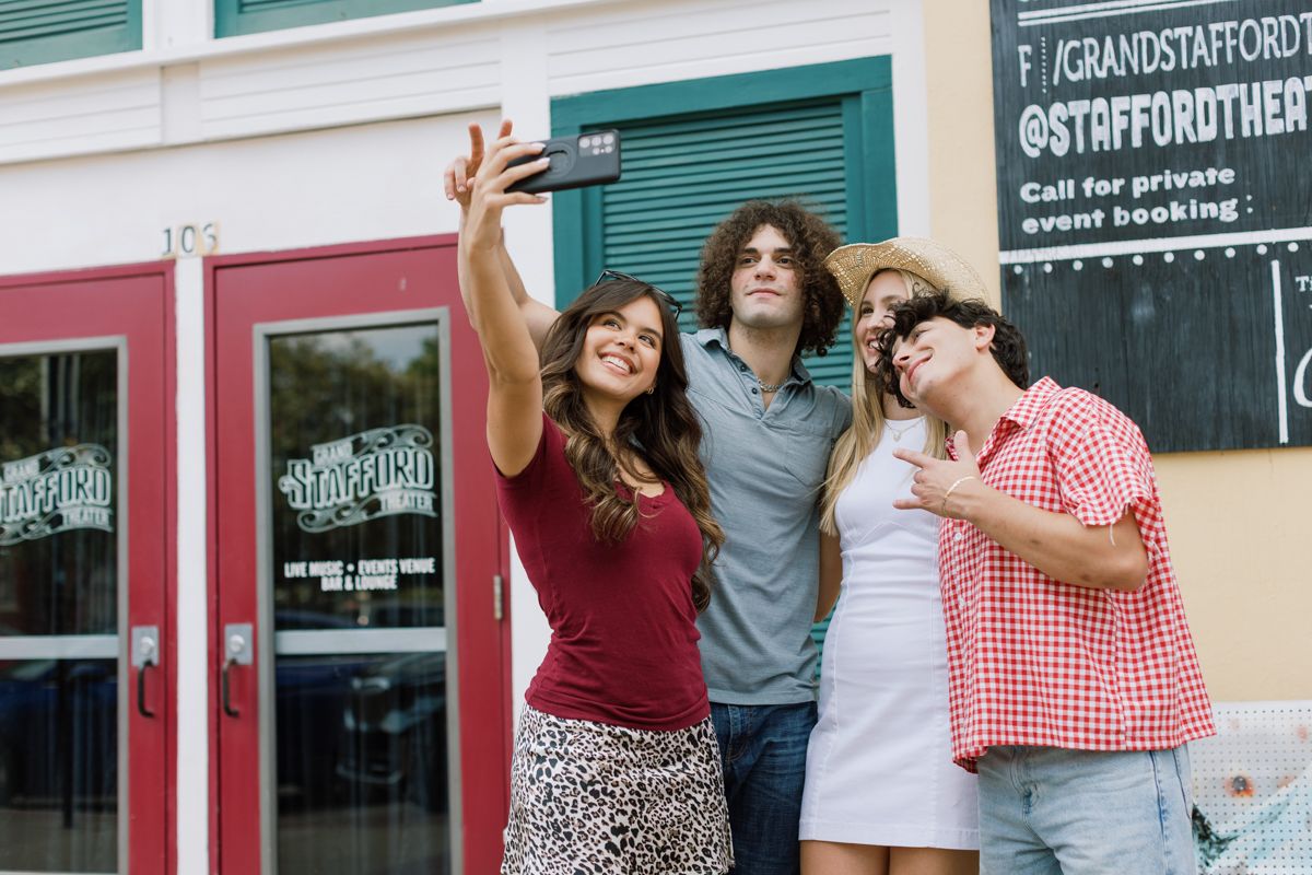 Four friends take a selfie outside the Stafford Theatre, smiling and posing together on a sunny day.