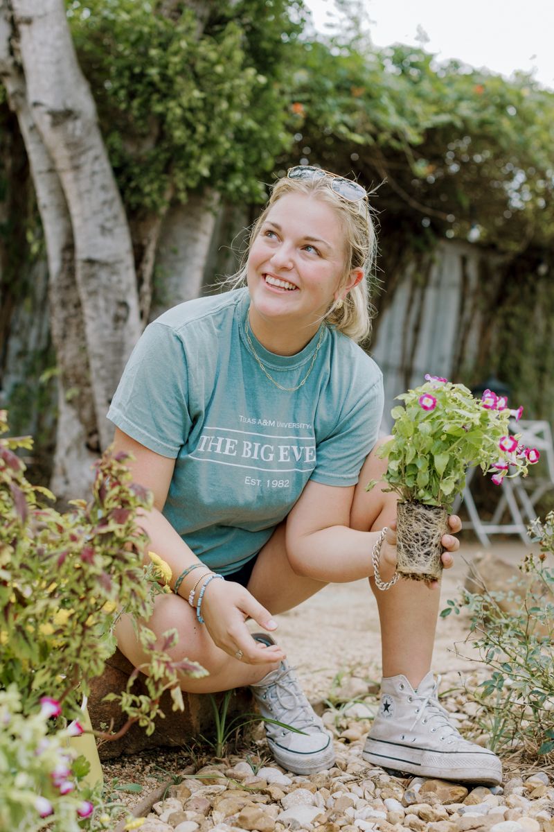 Smiling young woman crouching in garden holding small flowering plant while gardening outdoors.